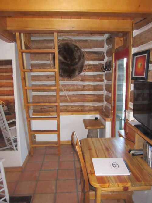 Cozy log cabin interior featuring a wooden ladder, table, and TV, with rustic walls and a round decorative piece.