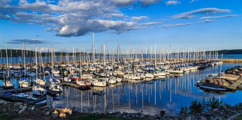 A scenic marina filled with sailboats, reflecting in calm water under a blue sky with scattered clouds.