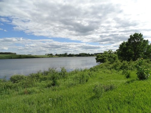 A serene landscape featuring a calm river, lush green grass, and trees under a partly cloudy sky.