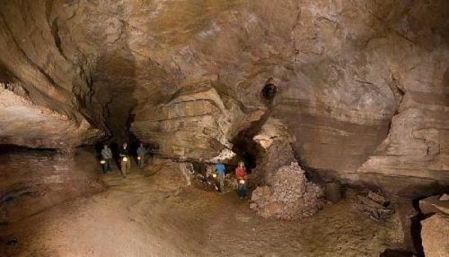 Explorers with headlamps navigate a large, rocky cave with uneven walls and a sandy floor.