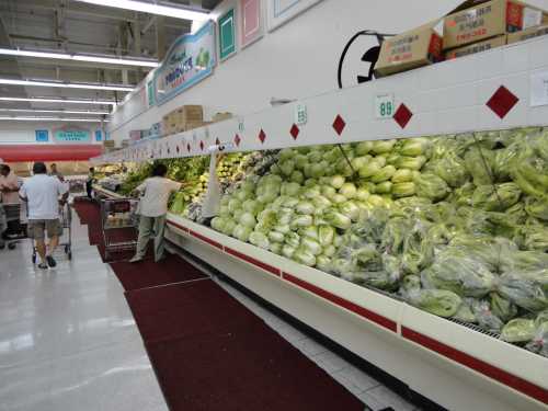 A grocery store aisle filled with various fresh vegetables, including cabbages and leafy greens, with shoppers nearby.