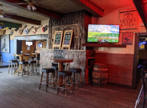 A rustic bar interior with wooden tables, stools, and a TV showing a sports game on the wall.