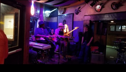 A live band performs in a cozy venue, featuring musicians with guitars and keyboards, surrounded by colorful decor.