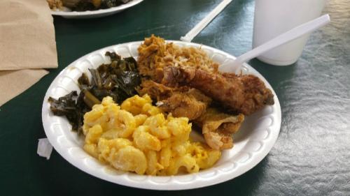 A paper plate with fried chicken, macaroni and cheese, collard greens, and rice, alongside a cup and a plastic spoon.