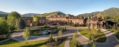 A scenic view of a large lodge surrounded by greenery and mountains, featuring a circular driveway and a pond.