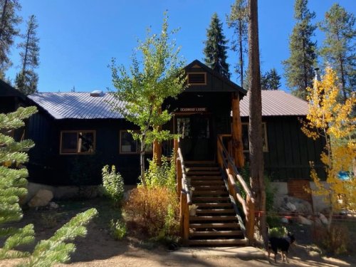 A rustic lodge with a wooden staircase, surrounded by trees and autumn foliage under a clear blue sky.