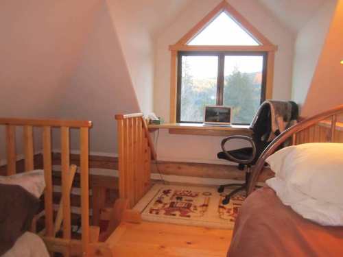 Cozy attic bedroom with a wooden railing, a window, and a small desk with a computer. Warm, inviting atmosphere.