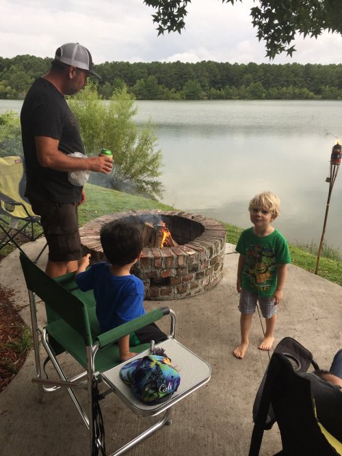 A man stands by a fire pit near a lake, while two children sit nearby, enjoying the outdoors on a cloudy day.