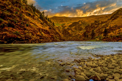 A serene river flows through a rocky landscape, surrounded by hills and trees under a colorful sunset sky.