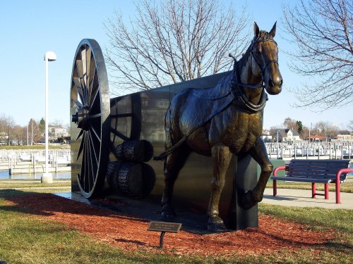 A bronze statue of a horse pulling a large wheel, set in a park near a waterfront.