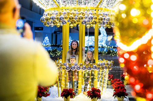 A woman and two children pose inside a festive, illuminated display with holiday decorations and lights.