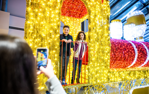 Two children smile on a brightly lit train display while a person takes their photo.