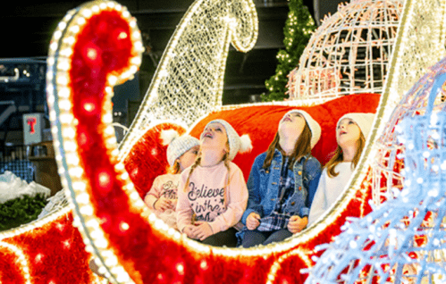 Four children in winter hats gaze up in wonder while sitting in a brightly lit, festive sleigh.