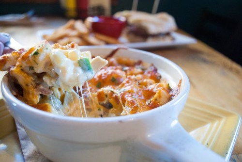 A creamy, cheesy dish being served from a white bowl, with melted cheese stretching and a plate of fries in the background.