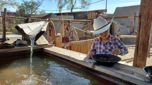 A child in a cowboy hat sifts through materials at a water trough in a rustic outdoor setting.