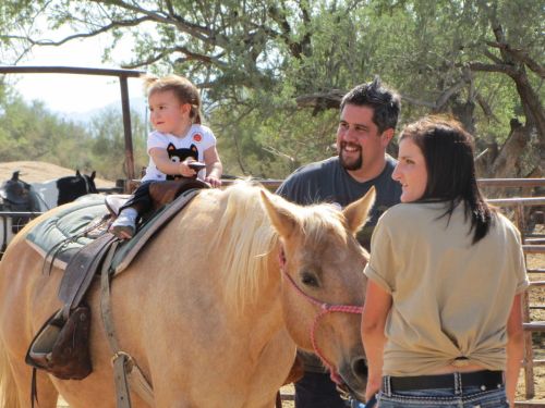 A smiling toddler sits on a horse, while two adults watch and smile in a sunny outdoor setting.