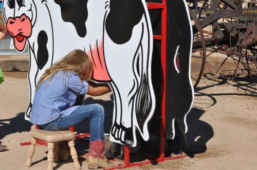 A child sits on a stool, pretending to milk a large, painted cow in a playful outdoor setting.