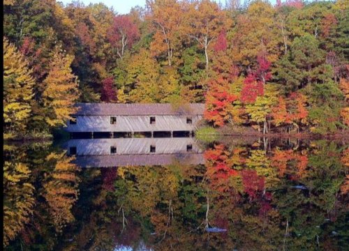 A rustic wooden building reflects in a calm lake, surrounded by vibrant autumn foliage in shades of red, orange, and yellow.