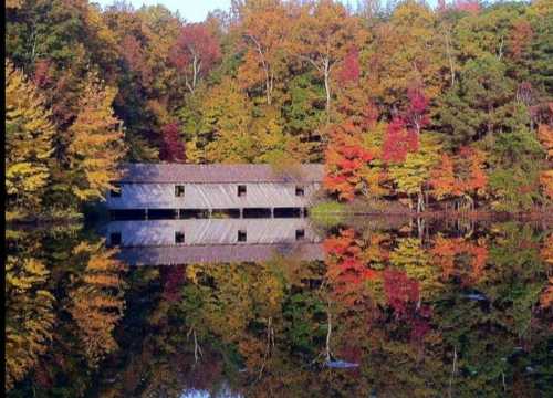 A rustic wooden building reflects in a calm lake, surrounded by vibrant autumn foliage in shades of red, orange, and yellow.