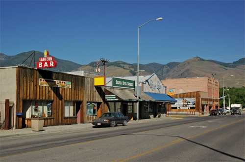 A small town street featuring rustic buildings, shops, and a bar, with mountains in the background under a clear blue sky.