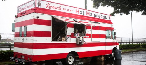 A red and white striped food truck named "Leonard's Bakery" selling hot malasadas, parked under a tree.