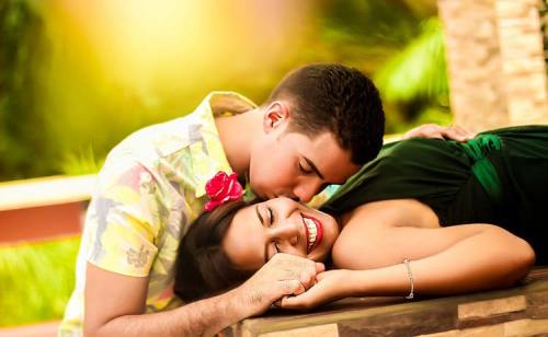 A couple shares a tender moment, smiling and embracing outdoors, surrounded by greenery and warm sunlight.