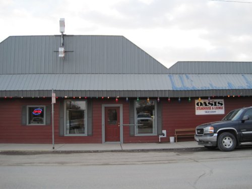 Exterior of a rustic restaurant named "Oasis Steakhouse & Lounge," featuring a gray roof and colorful lights.