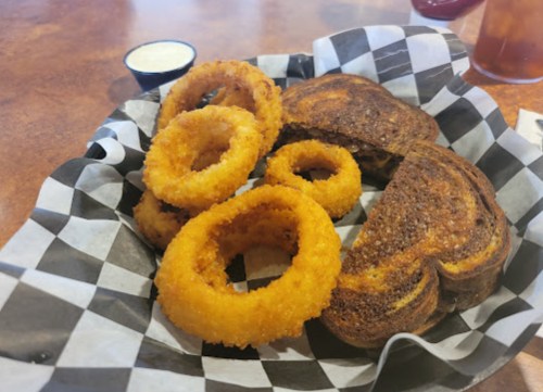 A basket with crispy onion rings and two slices of toasted bread on a checkered paper liner.
