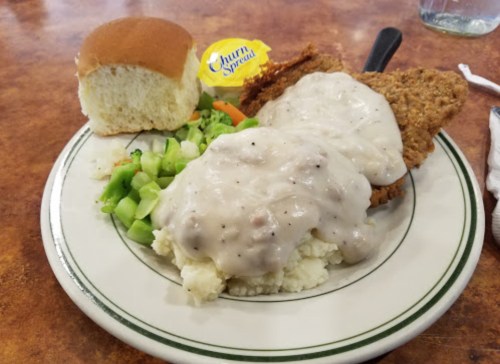 A plate with fried chicken, creamy gravy over mashed potatoes, a side of celery, and a roll.
