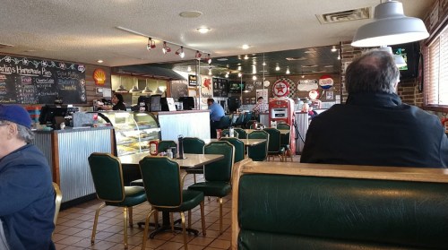 A cozy diner interior with green booths, tables, and a counter, featuring vintage decor and patrons enjoying their meals.