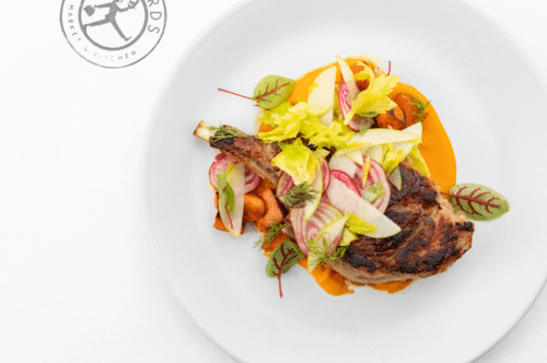 A beautifully plated pork chop with colorful vegetables and a vibrant sauce on a white dish.