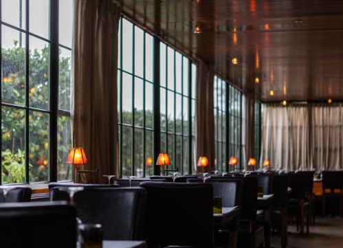 Cozy restaurant interior with dark chairs, warm lamps, and large windows overlooking greenery.