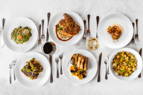A spread of six gourmet dishes on white plates, surrounded by silverware, with drinks in the background.