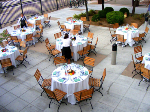 An outdoor dining area set with round tables, chairs, and neatly arranged tableware, with staff preparing for an event.