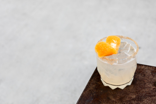 A refreshing cocktail with ice, garnished with two orange slices, sits on a wooden table against a light background.