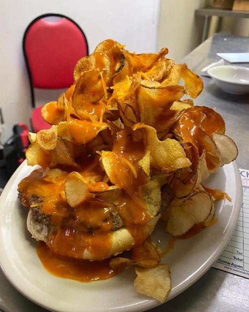 A large burger topped with crispy potato chips and drenched in orange sauce, served on a white plate.