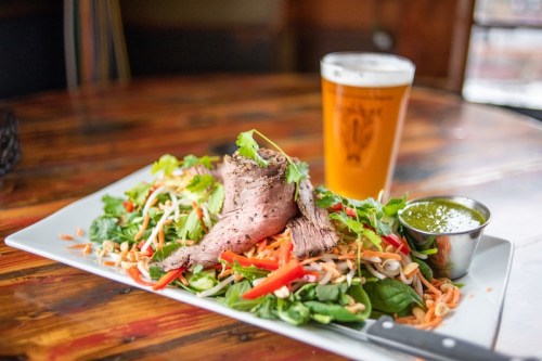 A colorful salad topped with sliced beef, served with a side of green sauce and a pint of beer.