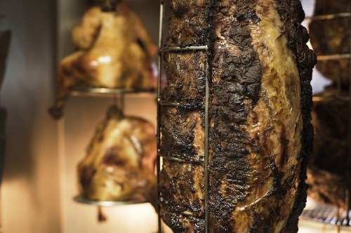 A close-up of a large, roasted meat cut on a spit, with additional meats visible in the background.
