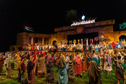 A large outdoor theatrical performance with actors in historical costumes, gathered on a stage at night.