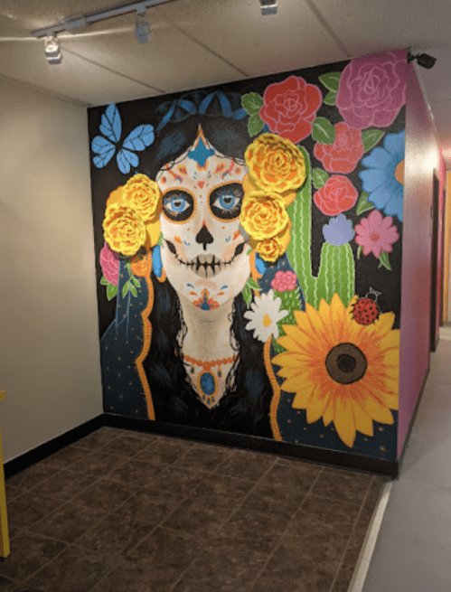 Colorful mural of a skull-faced woman surrounded by flowers, butterflies, and cacti on a black background.