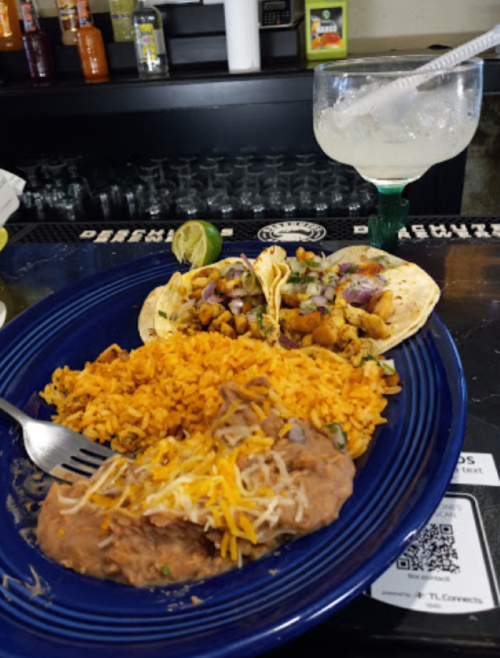 A blue plate with rice, refried beans, and two tacos, alongside a margarita on a bar counter.