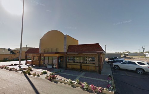 A colorful restaurant building with a curved roof, surrounded by flowers and parked cars on a sunny day.
