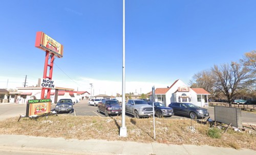 A roadside restaurant with a large sign reading "NOW OPEN," surrounded by parked cars and clear blue skies.
