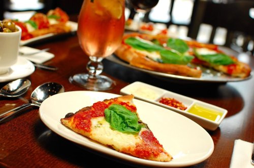 A slice of pizza topped with basil on a wooden table, accompanied by dipping sauces and a drink in the background.