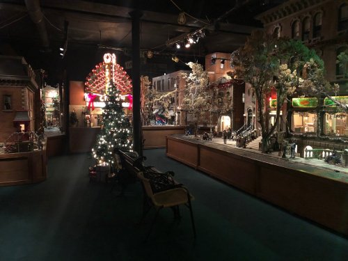 A dimly lit indoor scene featuring a decorated Christmas tree and a detailed miniature town with festive lights.