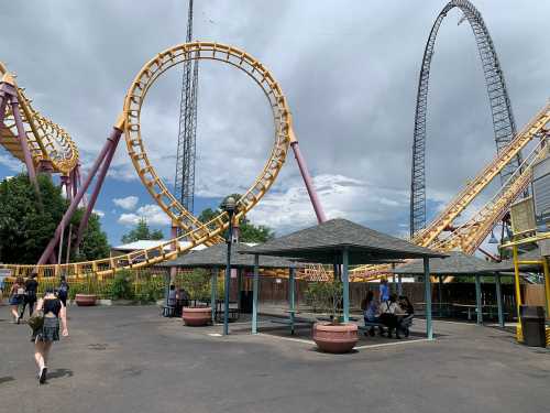 A roller coaster with loops towers over a park area with picnic tables and people walking nearby under a cloudy sky.