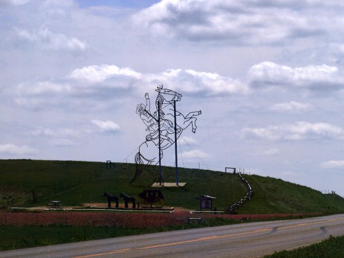 A large wire sculpture of a cowboy on horseback stands on a grassy hill under a cloudy sky, near a road.