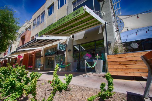 Exterior view of a modern restaurant with a green awning, surrounded by plants and outdoor seating. Bright blue sky above.
