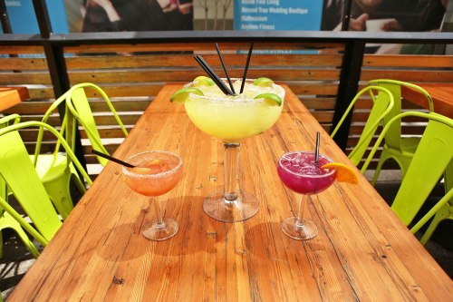 Three colorful cocktails on a wooden table, with a large margarita in the center and vibrant chairs in the background.