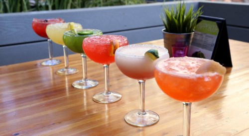 A row of colorful cocktails in large glasses on a wooden table, with a small plant and menu in the background.